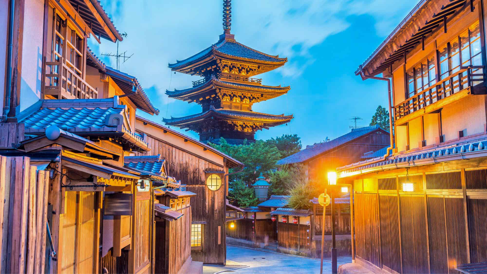 A traditional street in Kyoto features wooden houses with tiled rooftops, warmly lit by lanterns at dusk. The striking Yasaka Pagoda rises in the background, adding a timeless, cultural charm.