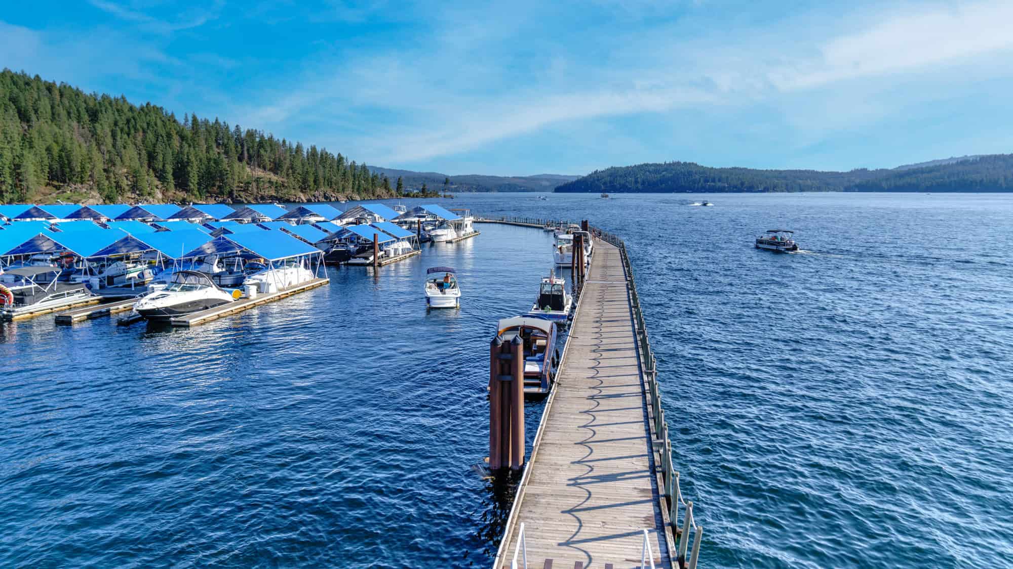 A long wooden boardwalk stretches over the sparkling blue waters of Lake Coeur d'Alene, surrounded by docked boats with bright blue canopies and forested hills in the distance.