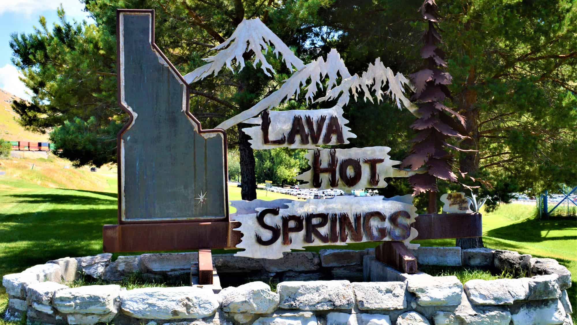 A metal cutout sign depicting snowy mountains, pine trees, and the shape of Idaho, with “Lava Hot Springs” in bold letters, nestled in a park-like setting.