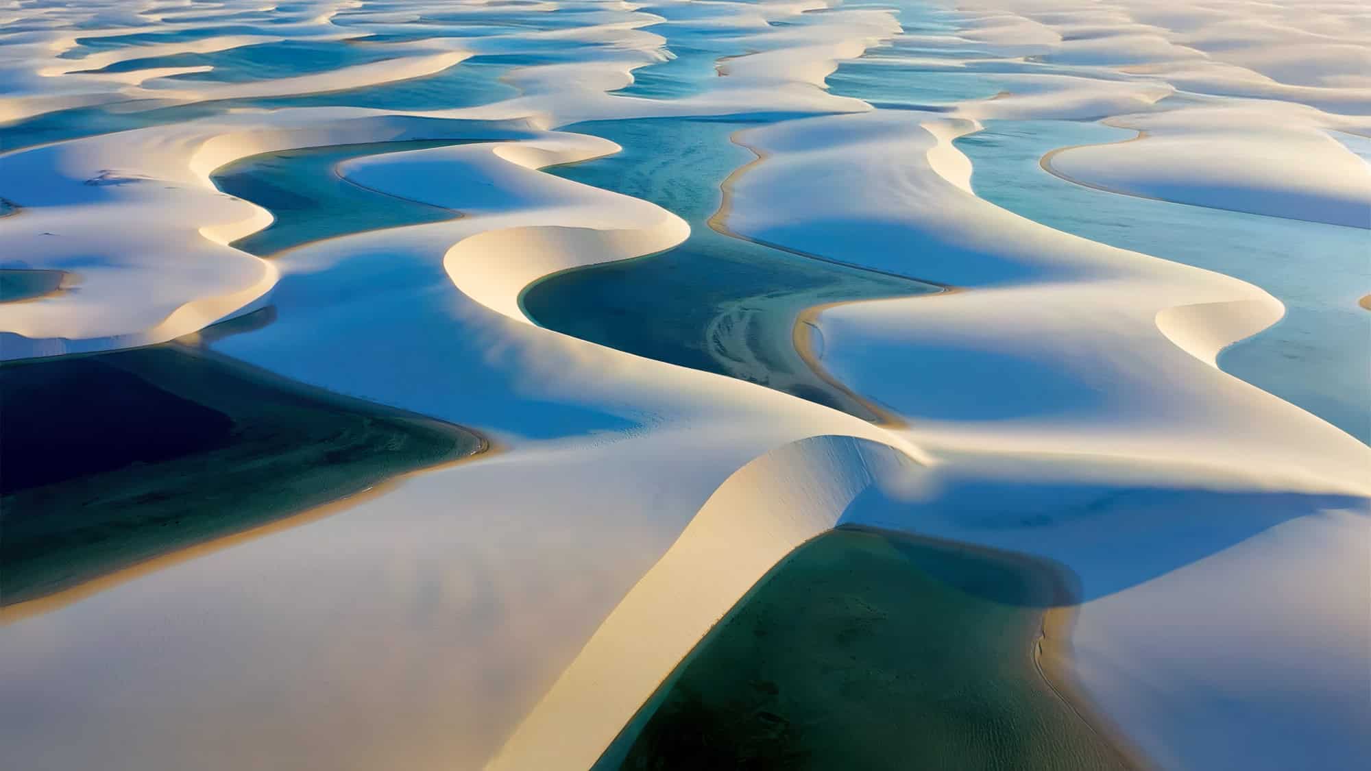 A surreal aerial view of Lençóis Maranhenses National Park in Brazil, showing undulating white sand dunes interspersed with turquoise and blue rainwater lagoons under soft lighting.