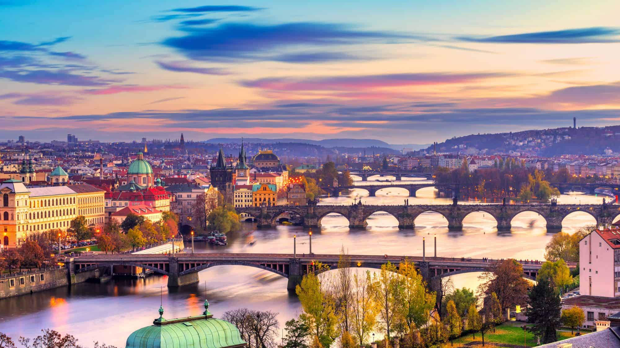Panoramic view of Prague from Letná Park, showcasing the Vltava River, multiple historic bridges including the Charles Bridge, and red-roofed buildings nestled among lush green trees.