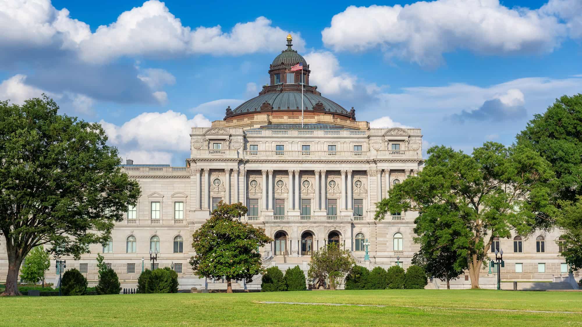 Elegant neoclassical building with a grand dome and ornate columns, framed by manicured lawns and leafy trees under a bright, cloudy sky.