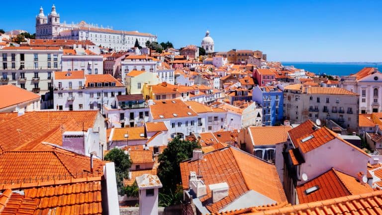 A panoramic view of Lisbon’s Alfama district shows a sea of orange-tiled rooftops, pastel buildings, and domes stretching toward the Tagus River under a cloudless blue sky.