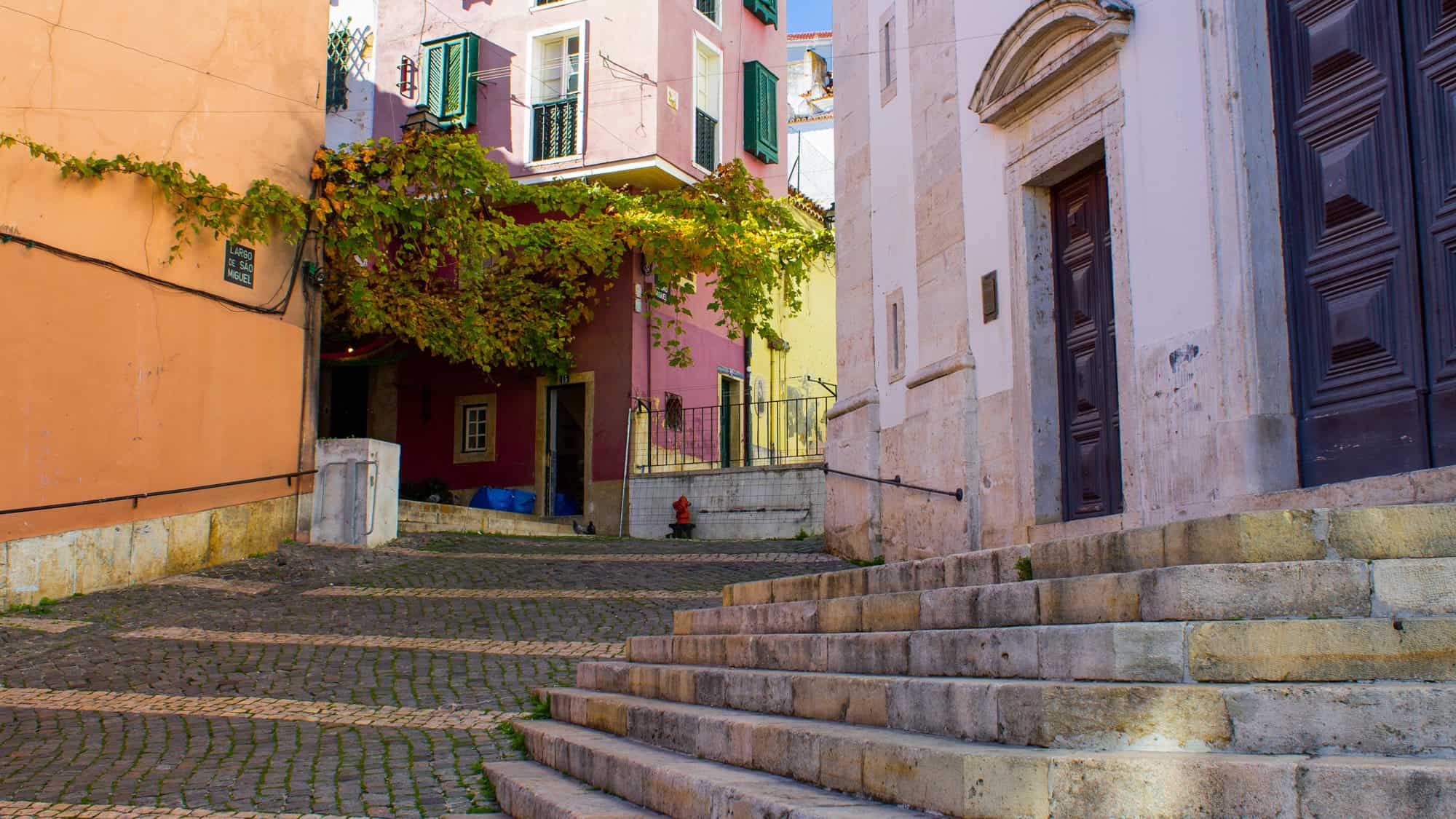 An uphill cobbled street in Lisbon winds past pastel-colored buildings with vines draping from balconies. A small staircase leads to a white stone church, and a red fire hydrant adds a splash of color.