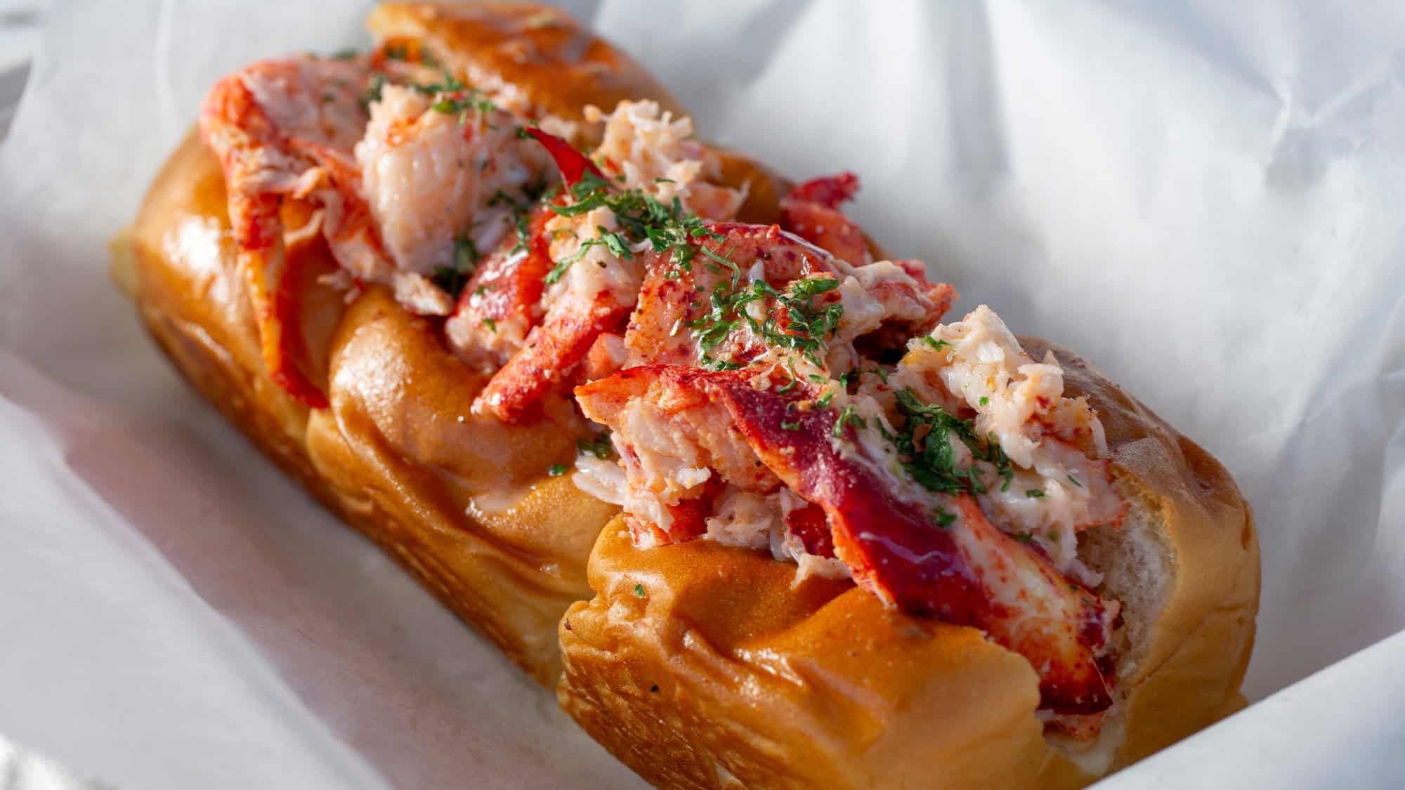 Close-up of a buttery, toasted split-top roll filled generously with fresh chunks of lobster meat, garnished with chopped herbs, and served on parchment paper.