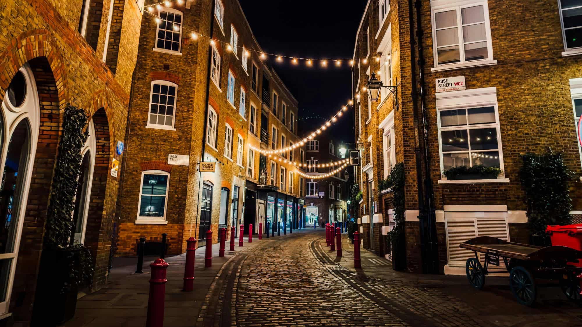 A narrow cobblestone street in London’s Soho district glows with strings of overhead lights. Red posts line the road, and historic brick buildings with white-framed windows create a cozy, intimate feel.