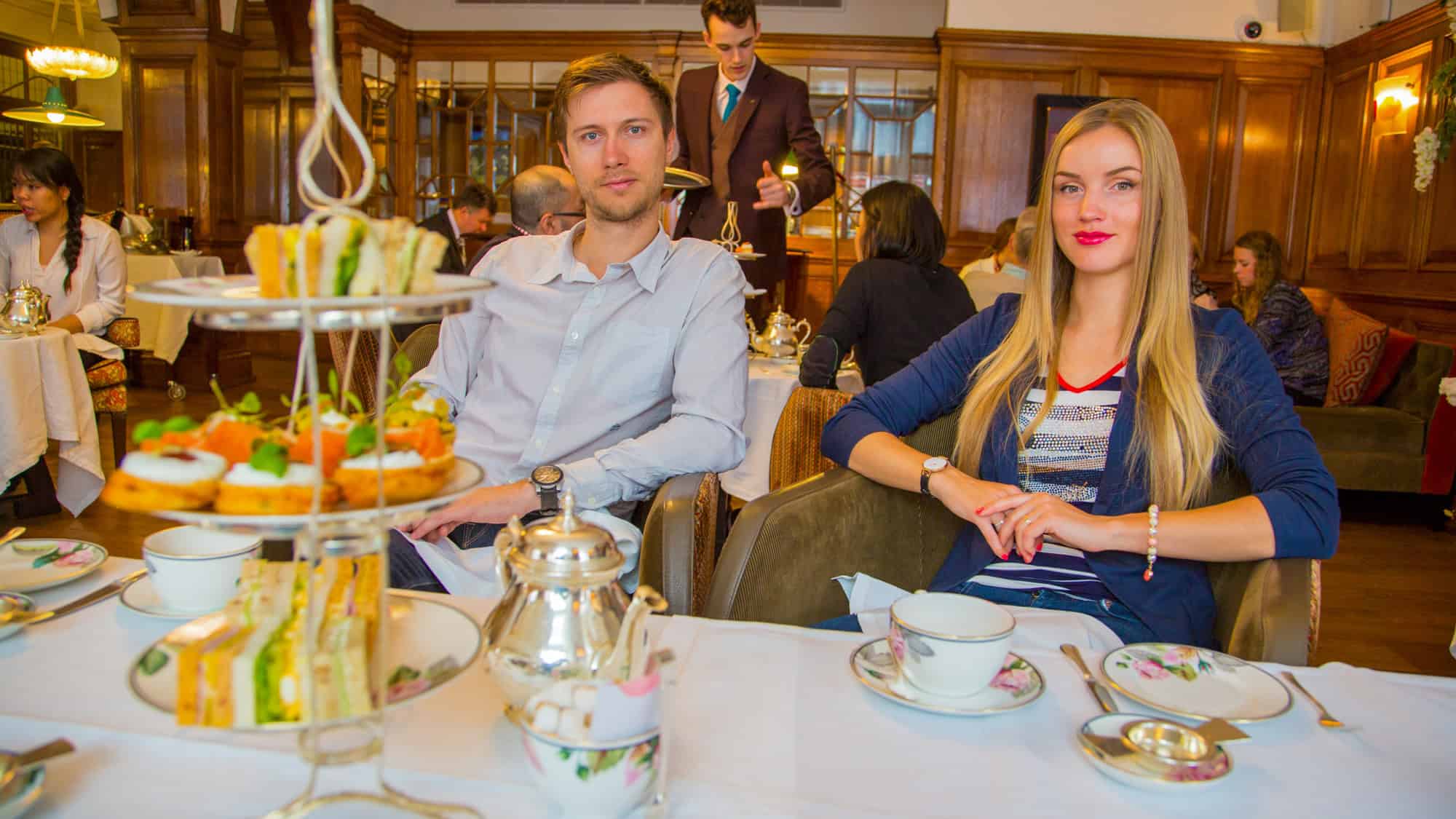 A man and woman sit at a table with a tiered tray of finger sandwiches and pastries in an elegant tea room.