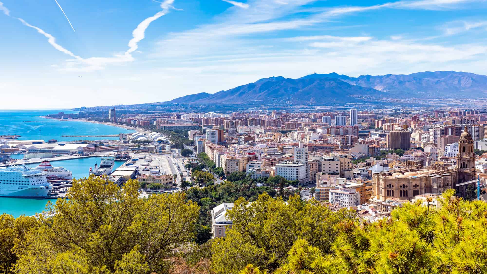 Overlooking Malaga, Spain, this cityscape features a busy harbor with cruise ships and piers, sprawling city blocks, historic architecture, and a backdrop of mountains and a vivid blue sky.