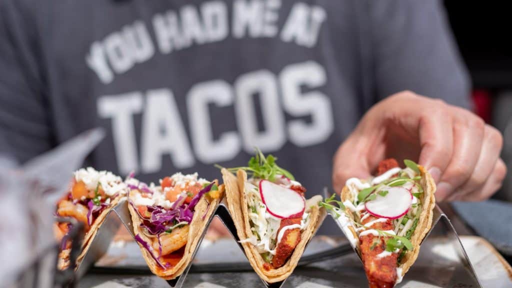 Three loaded tacos filled with shrimp, cabbage, radish, and fresh herbs are held in a metal taco stand, with a diner about to grab one.