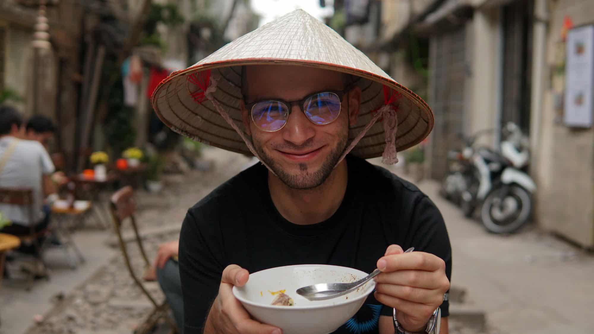A smiling man wearing a traditional conical hat enjoys a bowl of noodles at an outdoor street-side eatery.