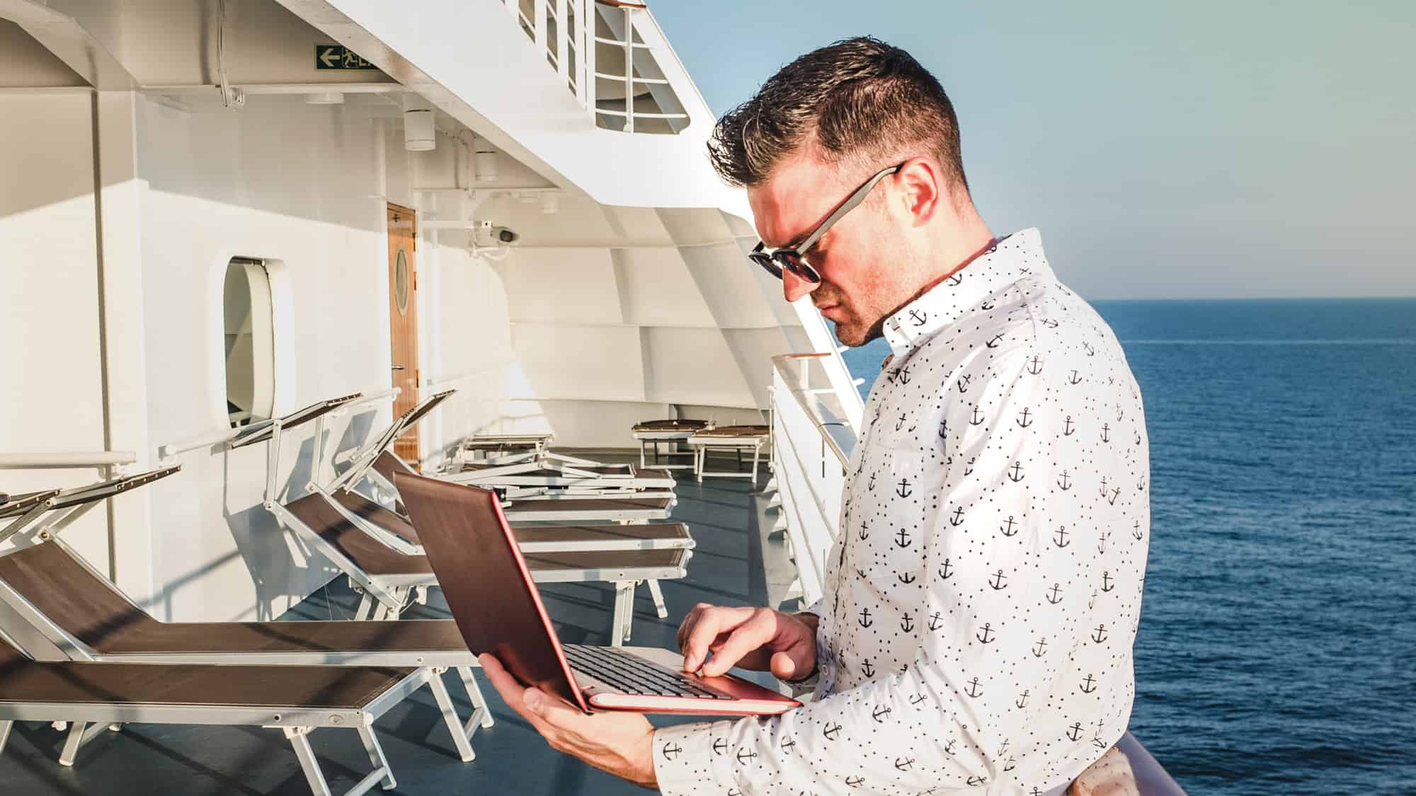 A man in a patterned white shirt and sunglasses types on a laptop while standing on a cruise ship deck, overlooking a calm, open sea.