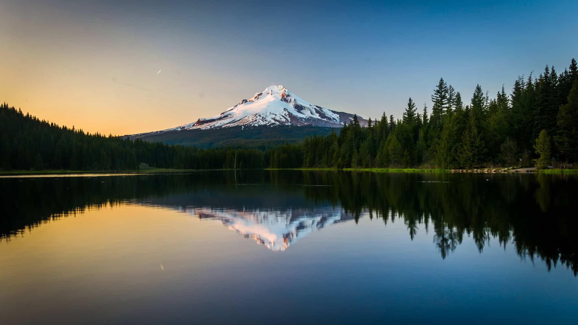A serene view of snow-capped Mount Hood reflected perfectly in the calm waters of Trillium Lake at dusk, framed by dense pine trees under a soft golden sky.