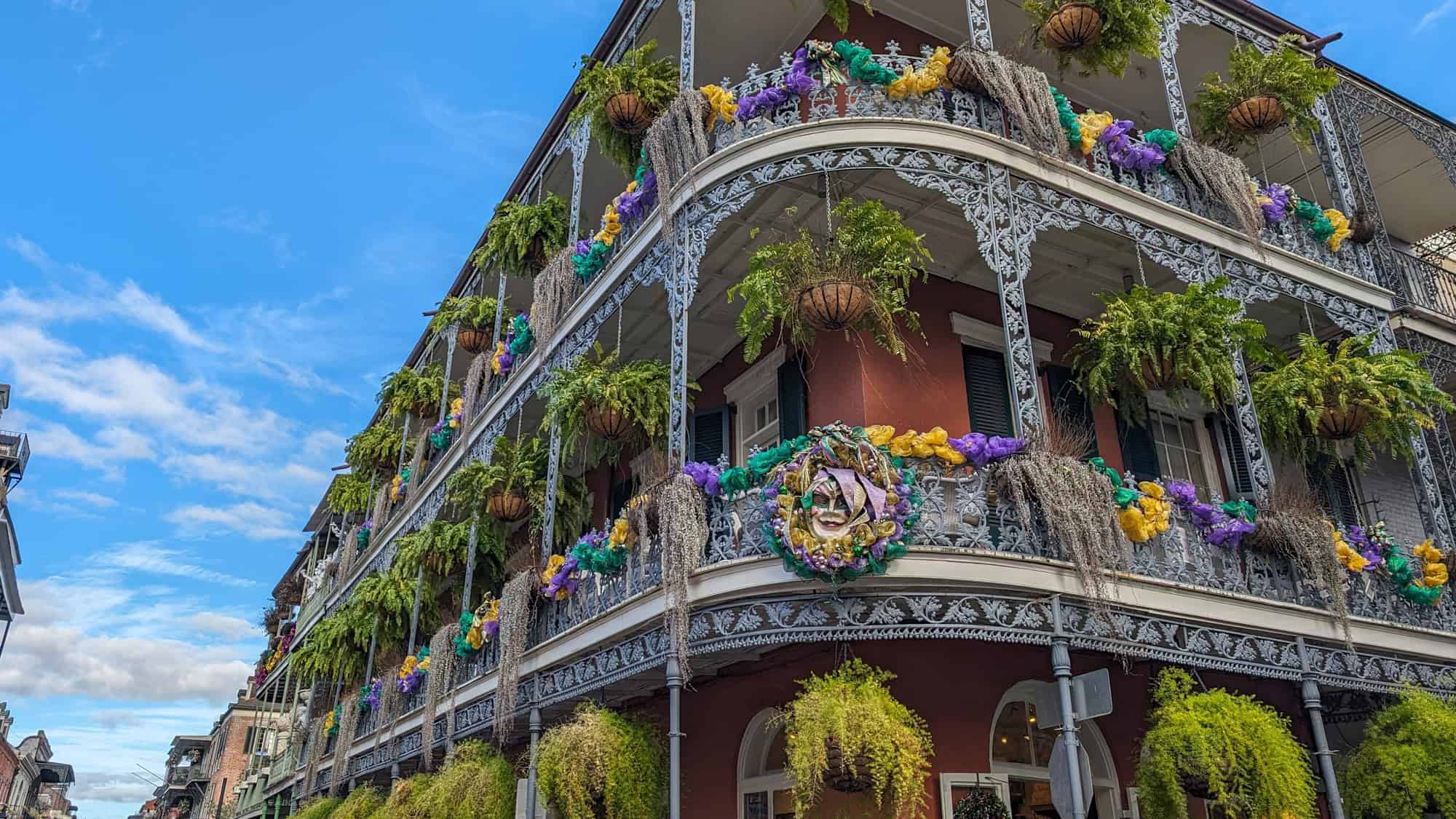 Colorful building with intricate wrought-iron balconies draped in Mardi Gras decorations and greenery, set against a bright blue sky.