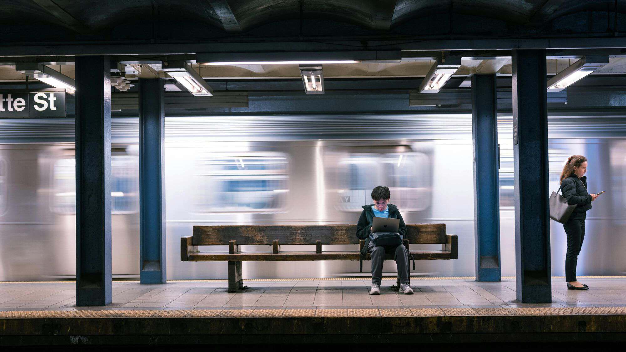 A man and a woman stand on a platform as a train whizzes by in a New York City subway station.
