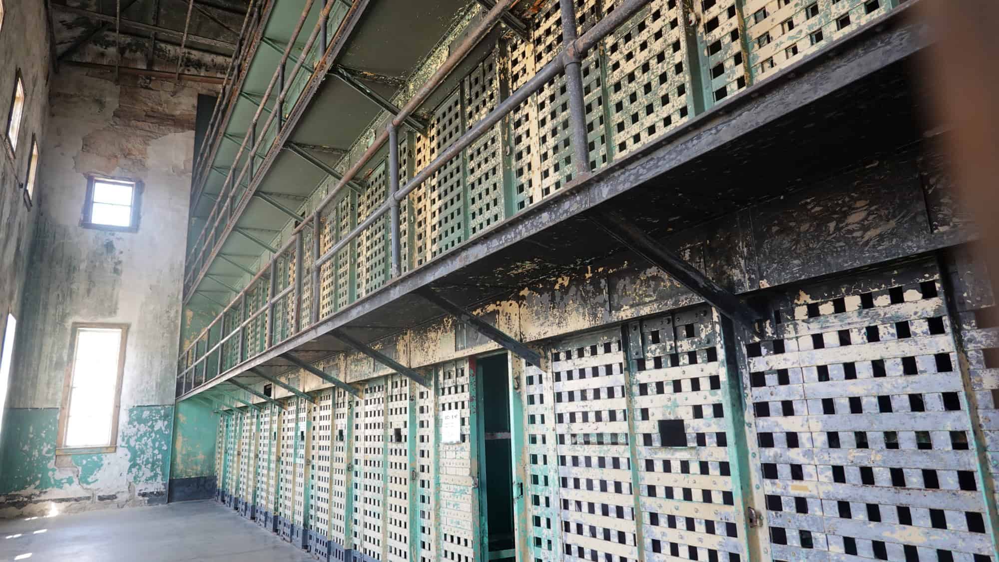 Inside a weathered, abandoned cell block of the Old Idaho Penitentiary, layers of peeling paint reveal the harsh past of this historic prison, with barred cells stretching across two levels.

