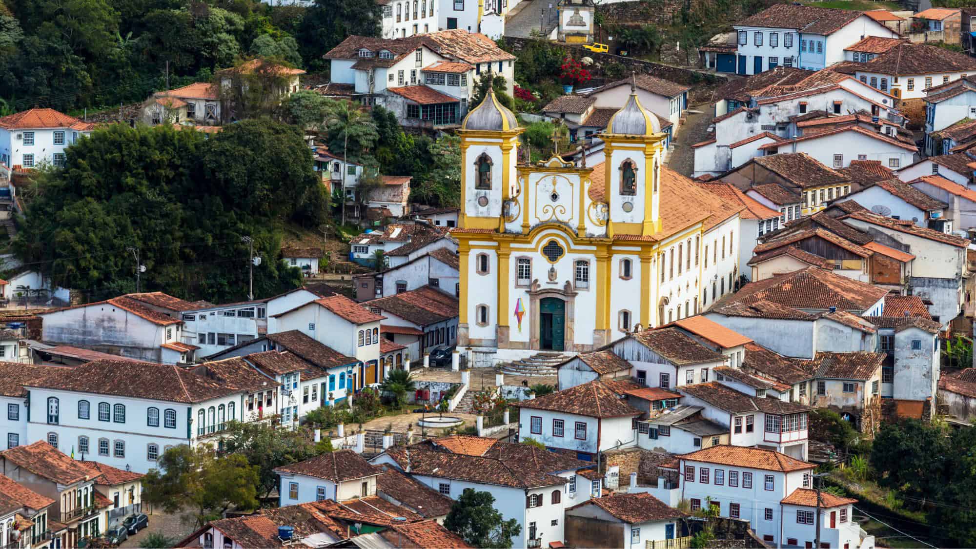 A colorful and compact colonial townscape in Ouro Preto, Brazil, with red-tiled rooftops, whitewashed houses, and a baroque-style church at the center of the frame.