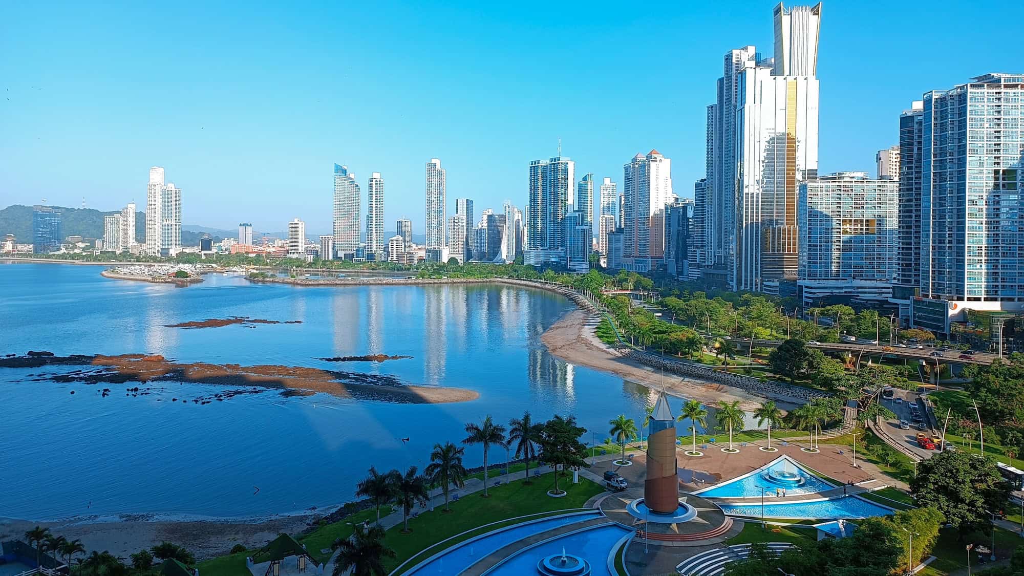 Bright and modern skyline of Panama City with tall skyscrapers reflecting in the bay, curving coastline, and palm-lined parks with blue pools and fountains in the foreground.