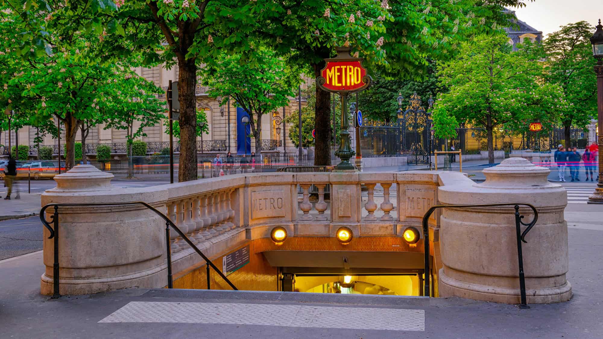 A classic Paris Metro entrance with ornate stonework and a red “METRO” sign, surrounded by lush green trees and elegant architecture in the background.