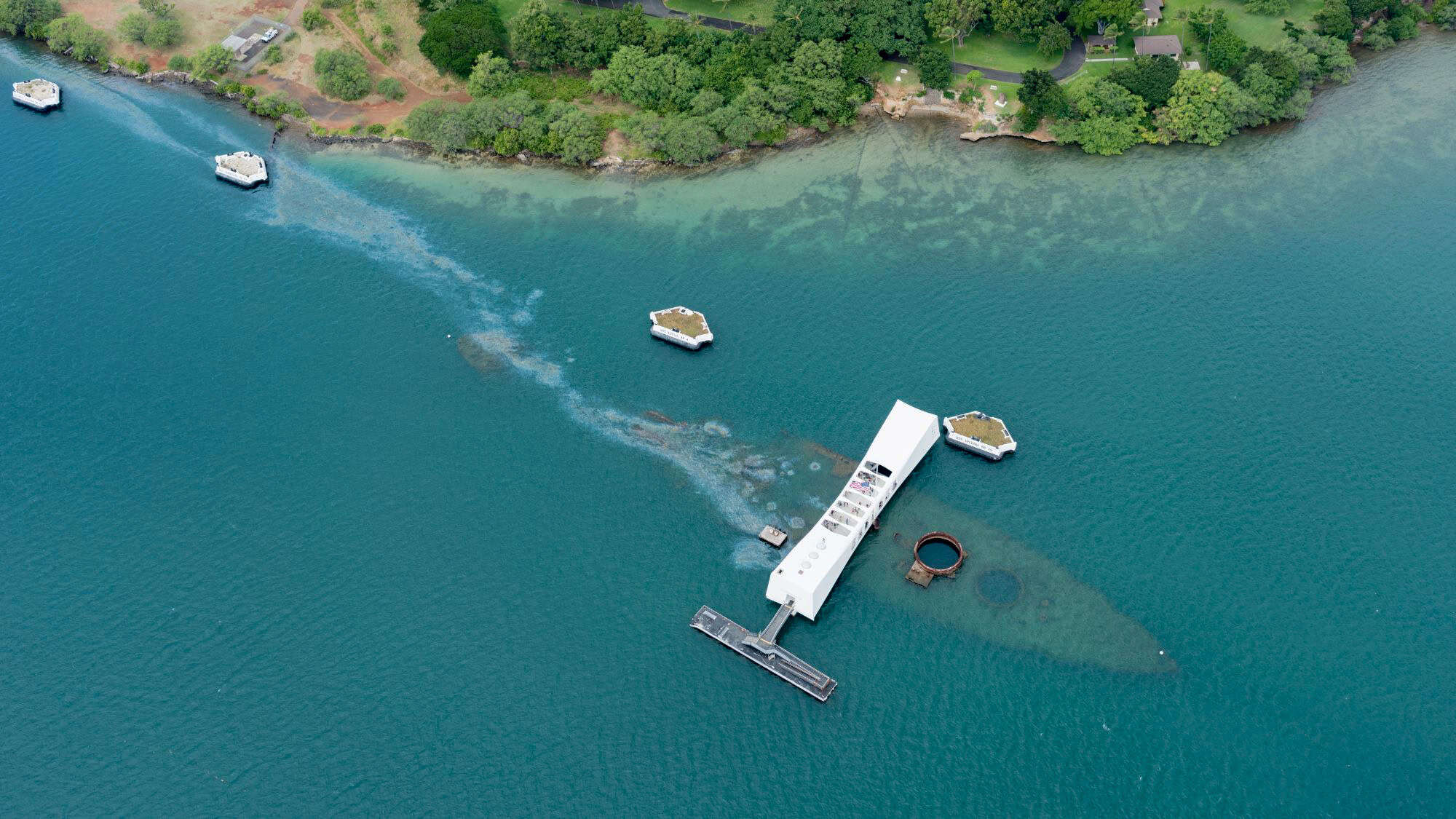 Aerial view of the USS Arizona Memorial, a white structure straddling the sunken ship visible beneath the teal waters of Pearl Harbor, with oil streaks still surfacing and green shoreline in the distance.