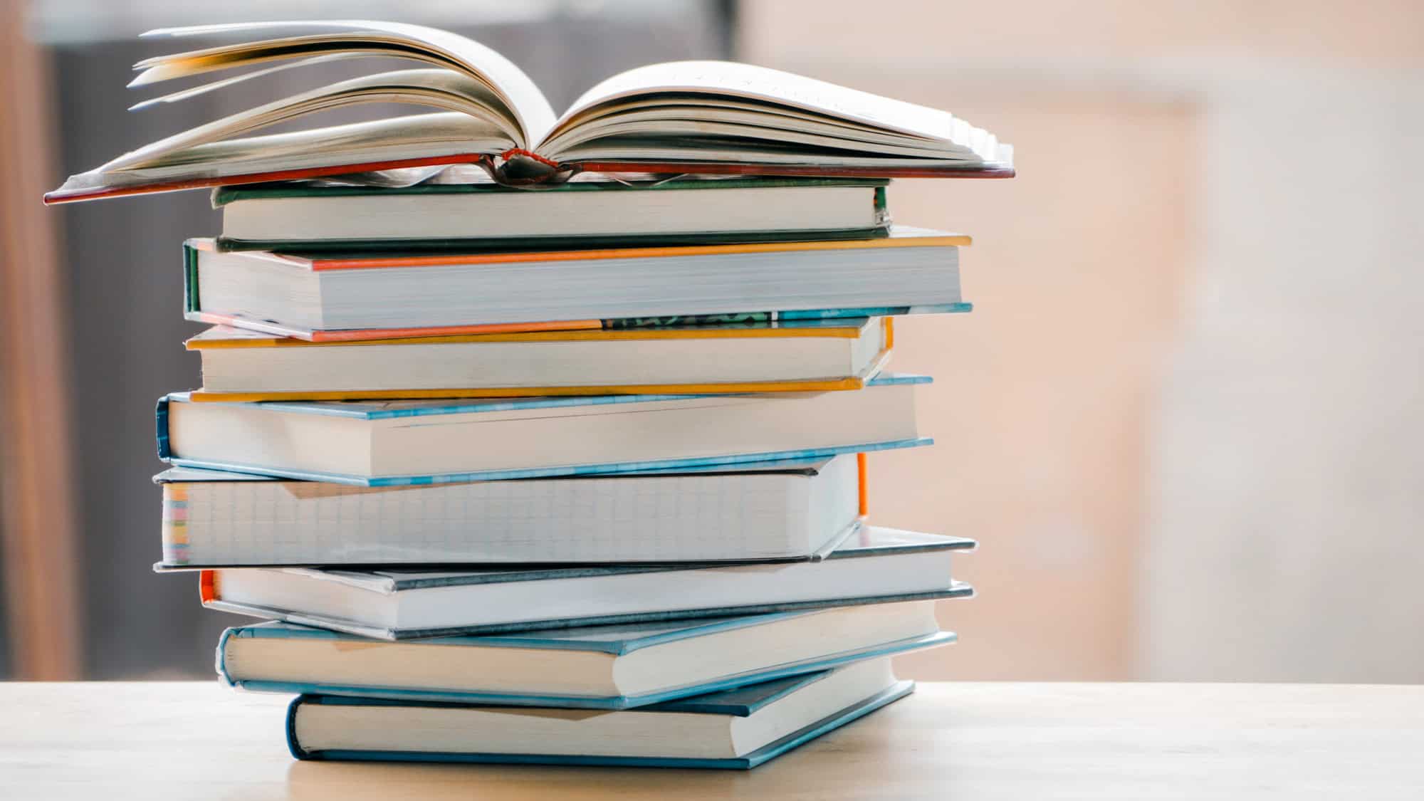 A tall, colorful stack of hardcover books sits on a light surface, with the top book open as if mid-read, creating a fan-like spread.