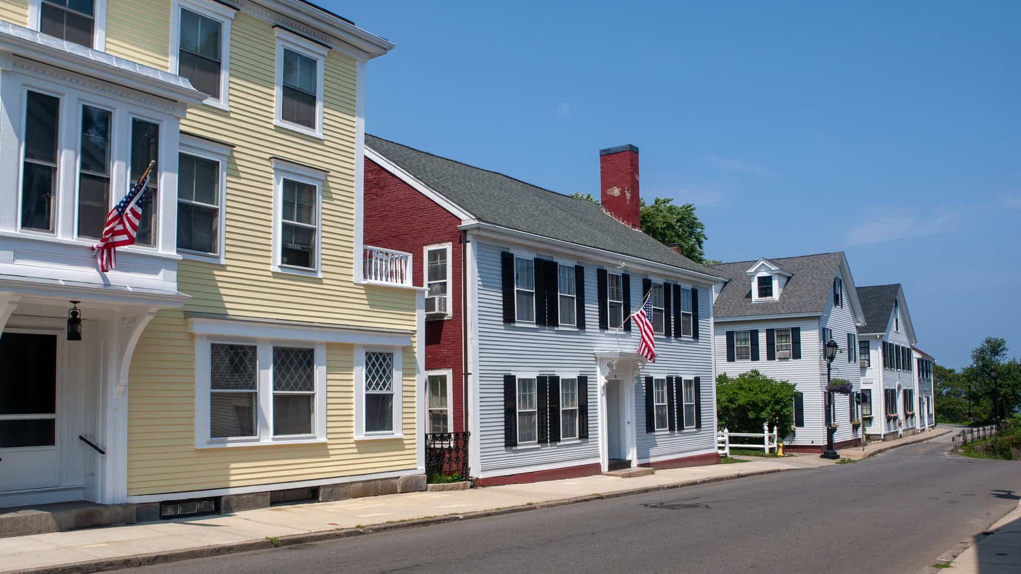 Quaint coastal New England street with colonial-style homes in pastel colors, many displaying American flags along a narrow road.