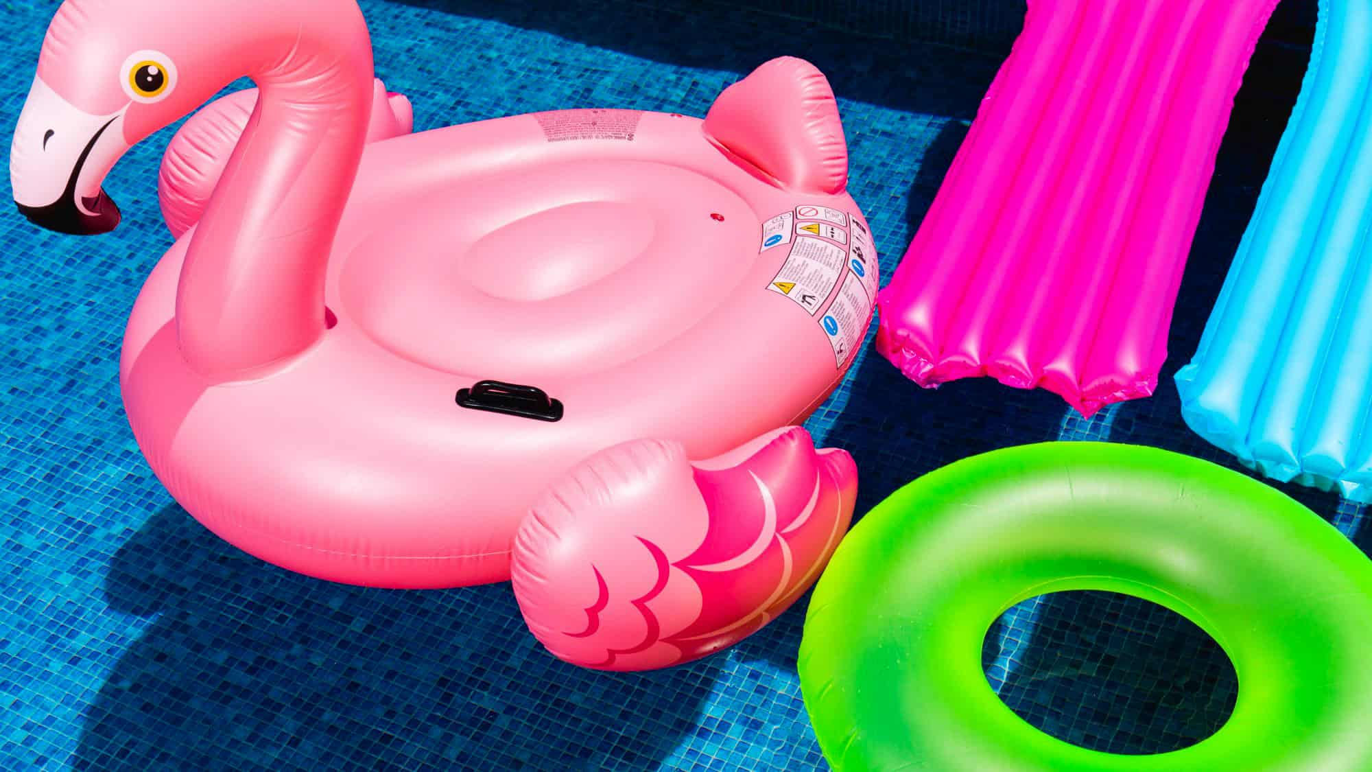 Bright inflatable pool floats, including a large pink flamingo, a neon green ring, and two air mattresses in pink and blue, float on a vibrant blue pool surface.