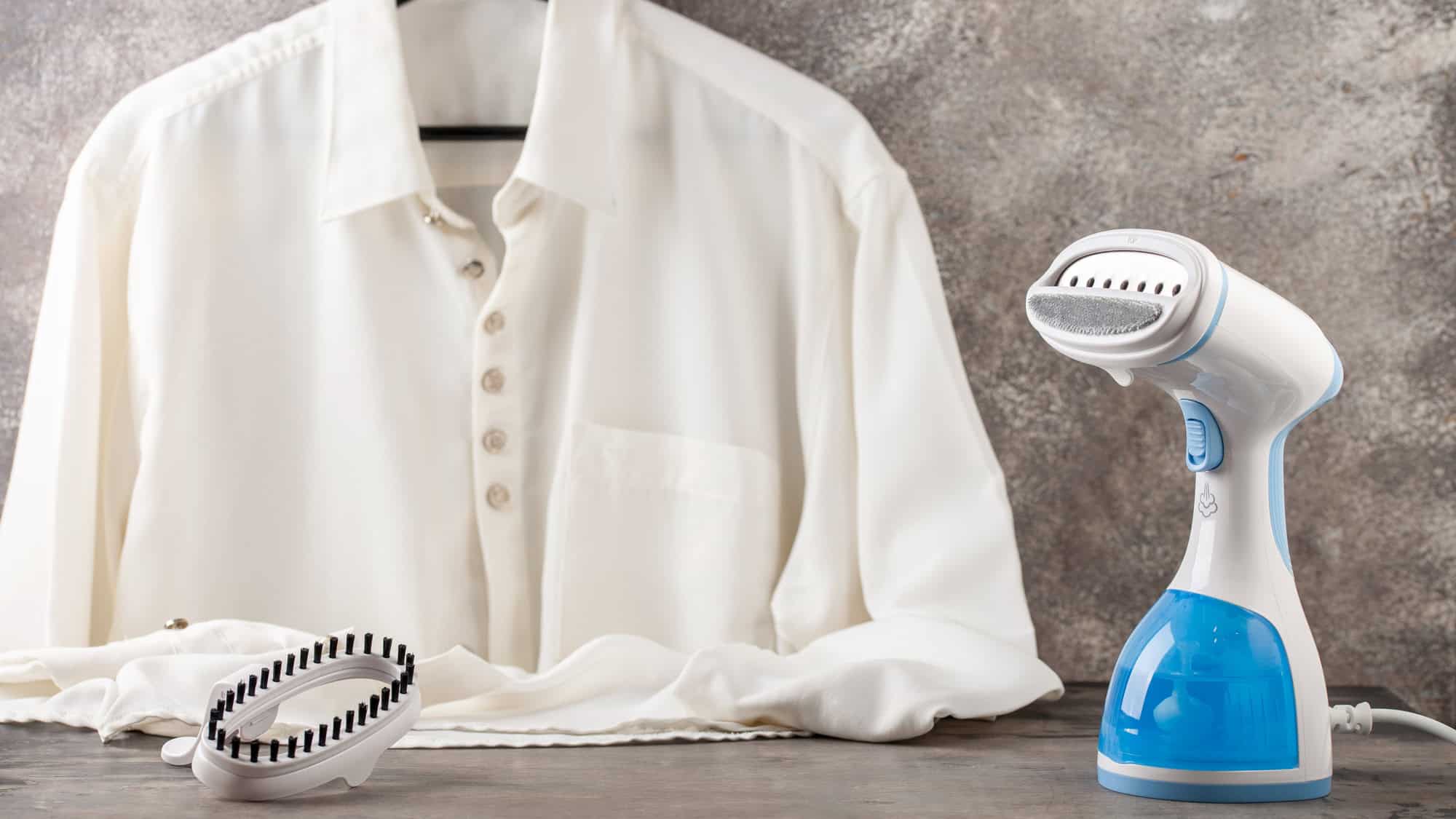 A white button-up shirt hangs on a hanger next to a portable garment steamer with a blue water tank, ready for use, resting on a countertop with a neutral textured backdrop.