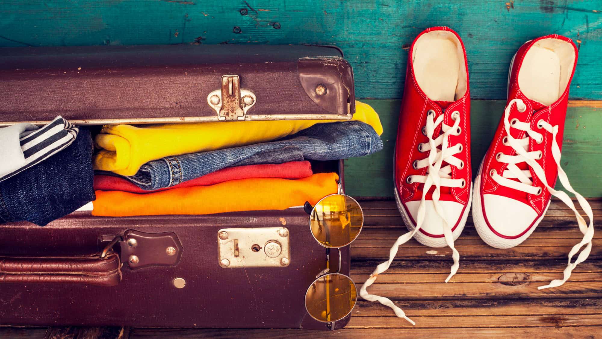 A vintage brown suitcase sits open, packed with rolled colorful clothes in yellow, red, orange, and denim, with a pair of bright red sneakers and aviator sunglasses placed beside it on a rustic wood floor.