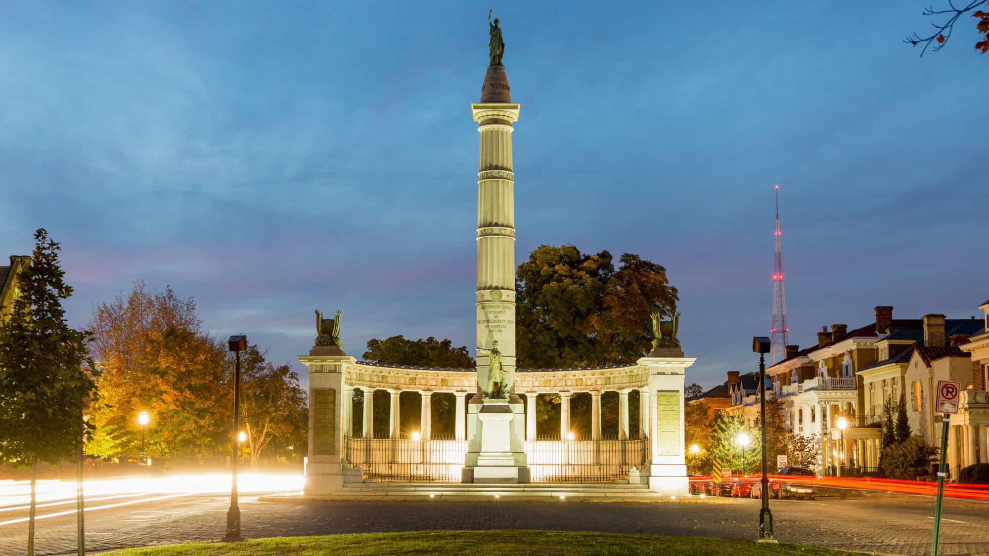 A tall, neoclassical-style monument with a bronze statue atop a fluted column, surrounded by a curved colonnade and glowing warmly against a twilight sky, set in the historic district of Richmond.