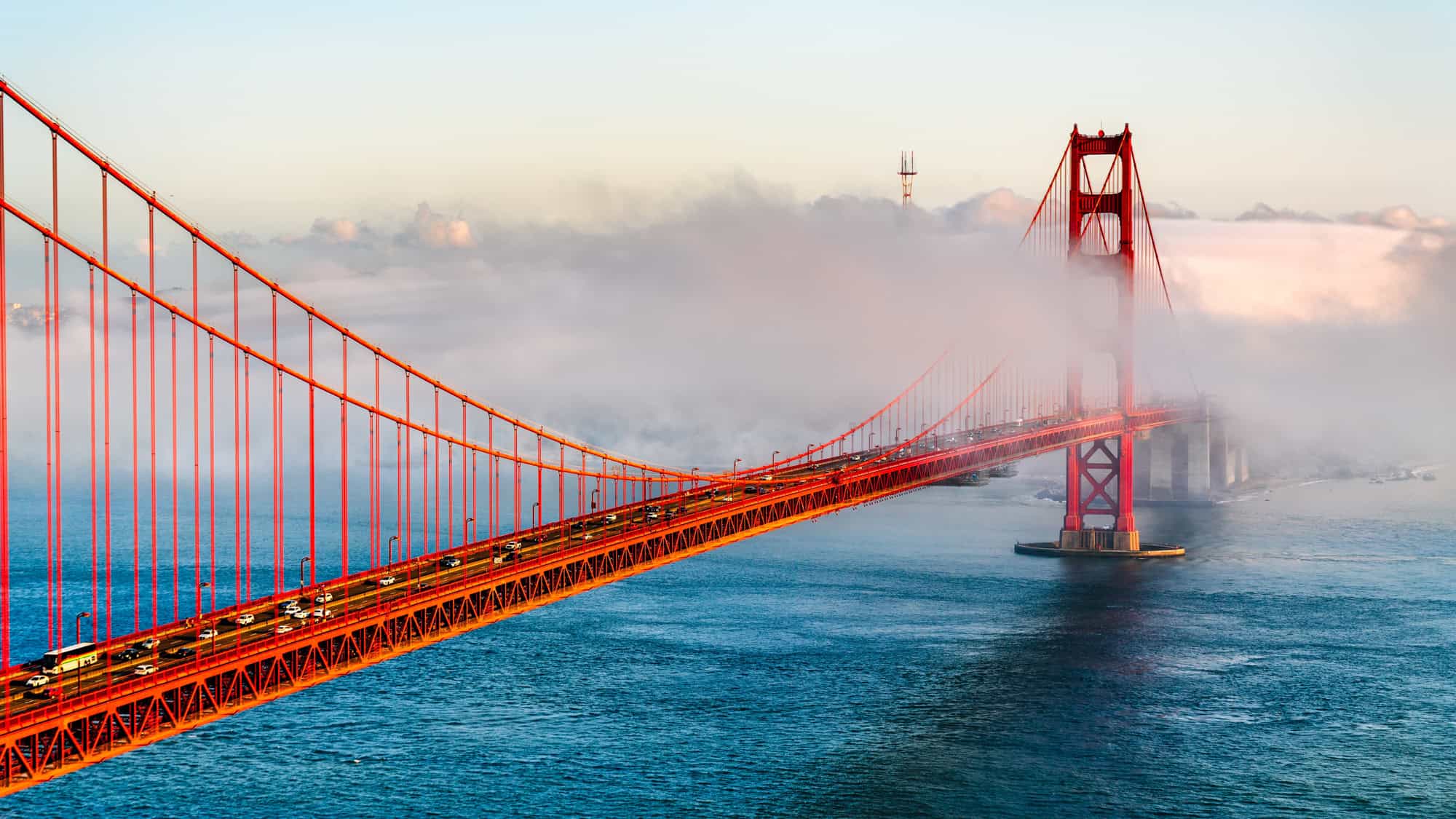 The Golden Gate Bridge stretches through dense fog over blue waters, with its iconic red towers emerging dramatically as cars cross the span beneath a pastel sky.