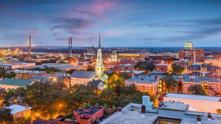 A sweeping view of Savannah, Georgia at twilight with glowing city lights, historic churches and buildings, and the Talmadge Memorial Bridge in the distance, all under a softly streaked evening sky.
