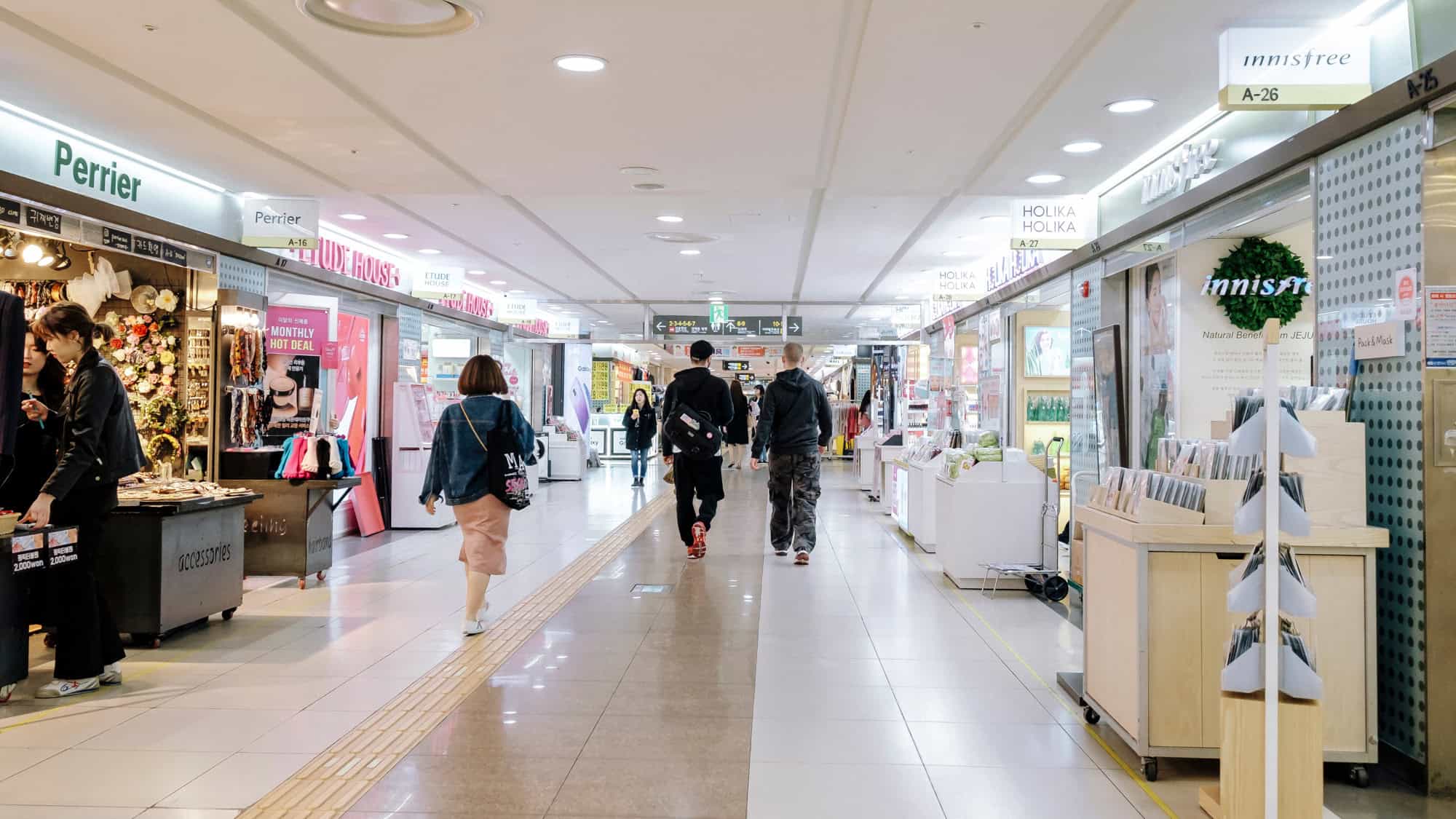 A sleek underground shopping mall in Seoul, lined with bright signage from Korean beauty and fashion brands, with shoppers walking past well-lit storefronts.