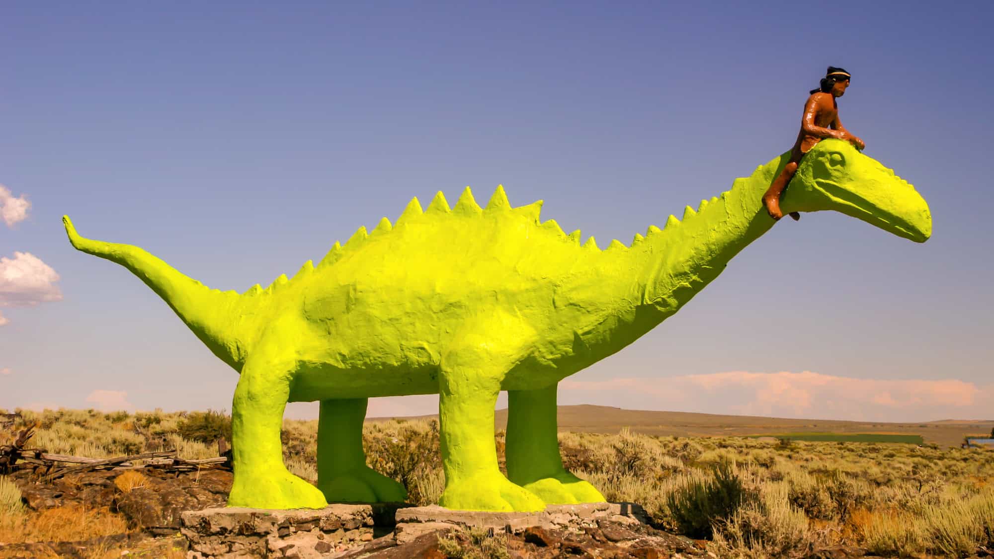 A bright green dinosaur sculpture stands in a desert landscape under clear skies, with a small Native American figure perched on its neck—an eye-catching piece of roadside Americana.