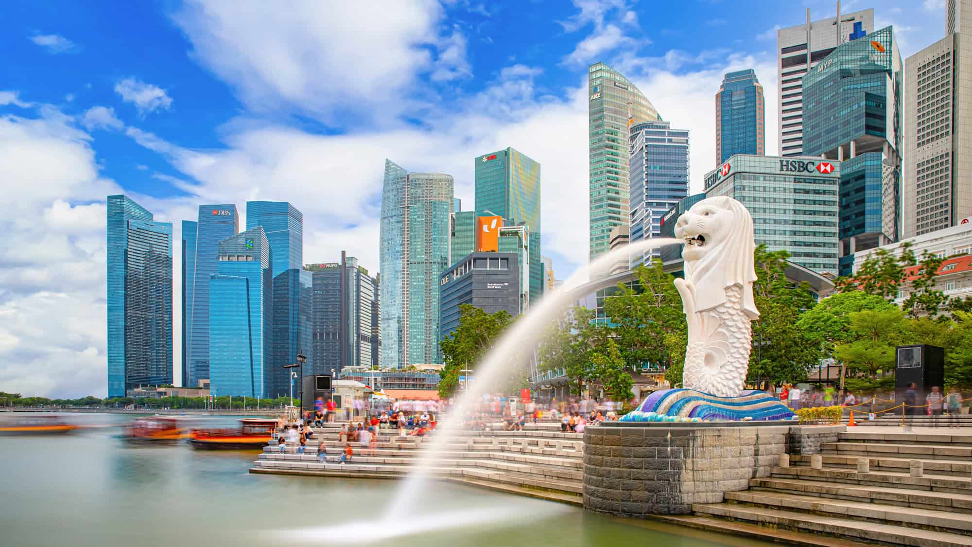 The famous Merlion statue in Singapore spouting water into Marina Bay, with a crowd gathered on tiered steps and sleek skyscrapers towering in the background under a vibrant blue sky.