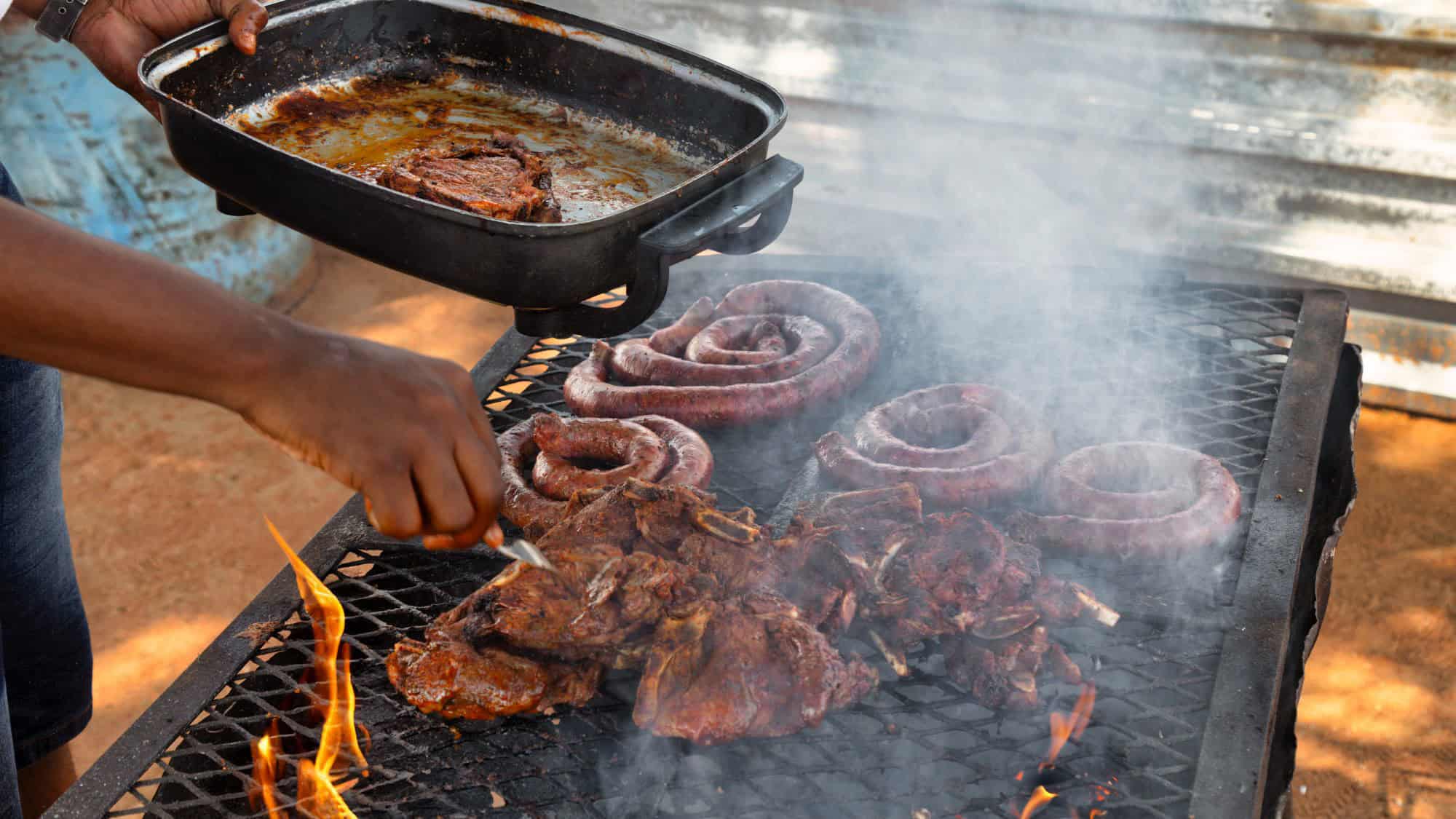 Coiled sausages and marinated meat sizzle over an open flame as a cook bastes them with sauce on a smoky grill.