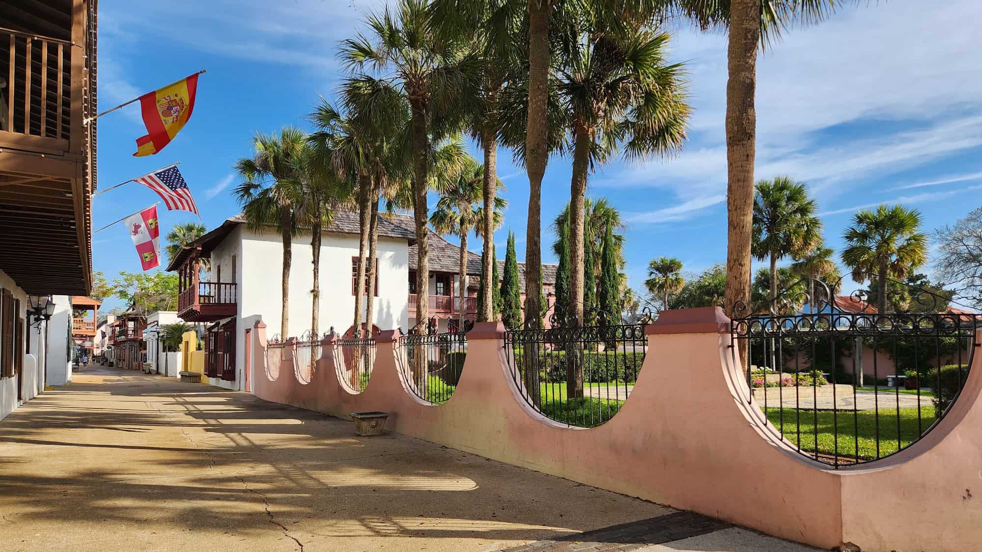 Charming colonial-style street lined with palm trees and historic buildings, including balconies and flags of Spain, the U.S., and Florida.