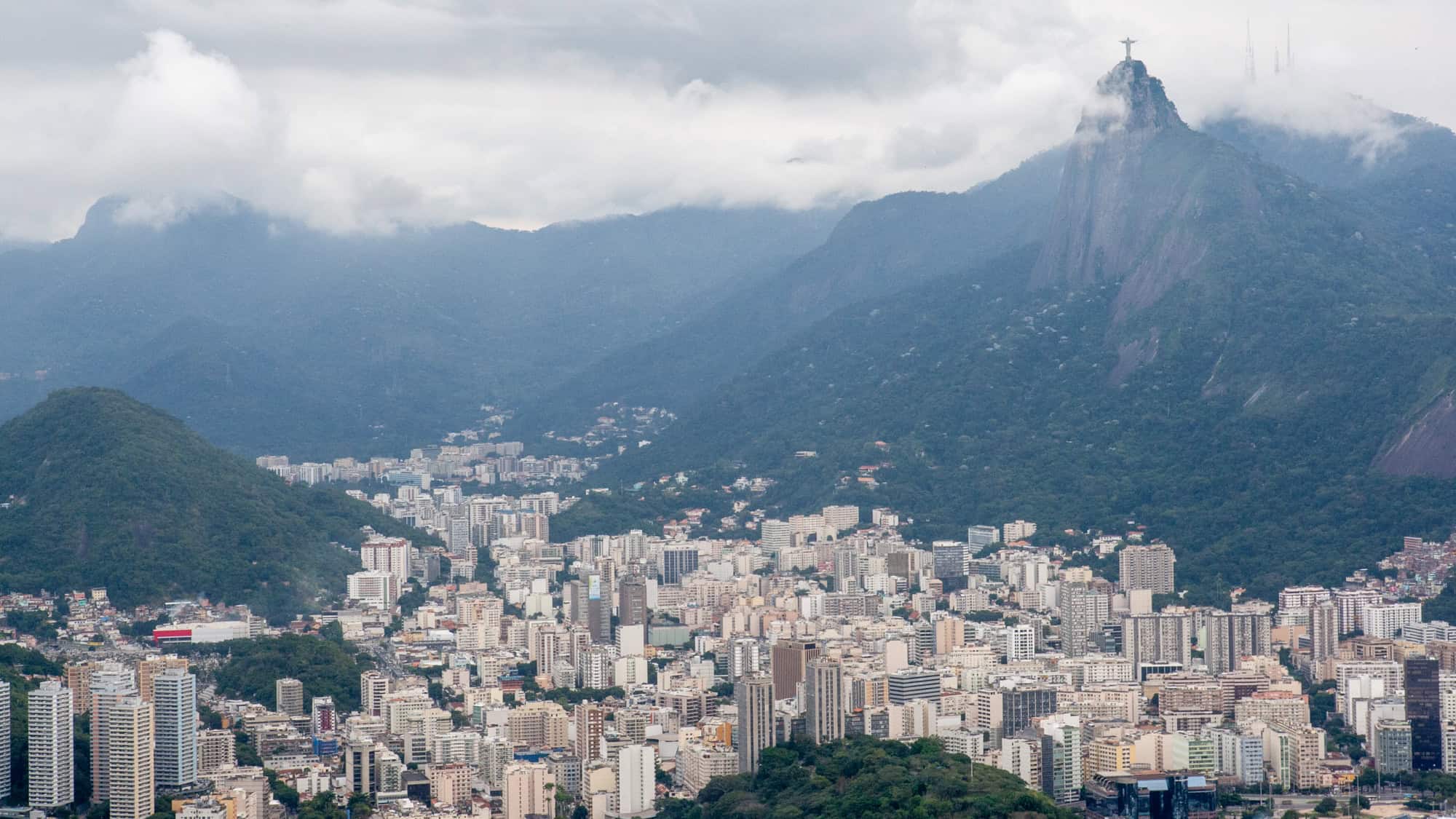 A cloudy view over Rio de Janeiro’s cityscape, with the iconic Christ the Redeemer statue emerging atop Corcovado Mountain, nestled among misty green peaks.