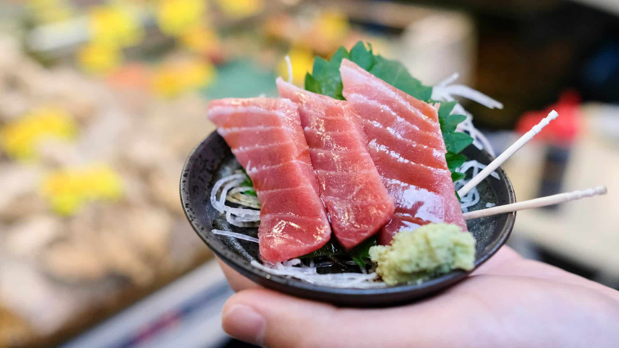 A small black bowl holds three slices of fresh tuna sashimi on shredded daikon with wasabi and shiso leaf.
