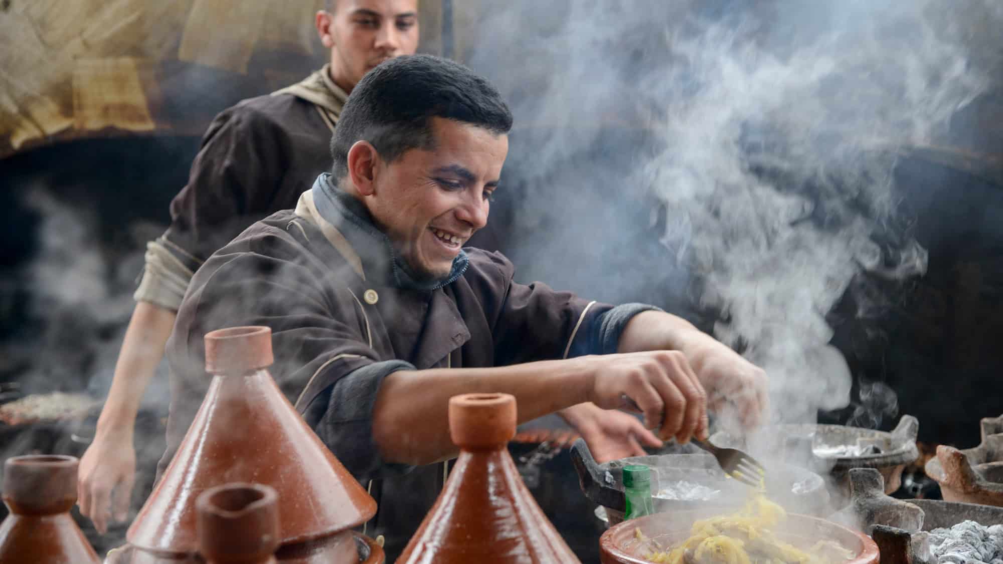 A smiling chef stirs steaming food in a clay tagine pot, surrounded by other tagines in a busy kitchen.