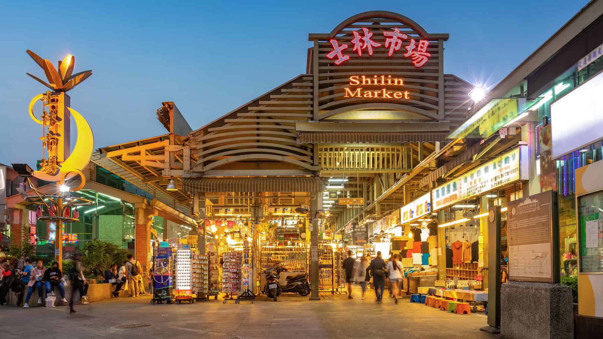 The brightly lit entrance to Shilin Market in Taipei glows at dusk, with stalls and crowds visible inside.