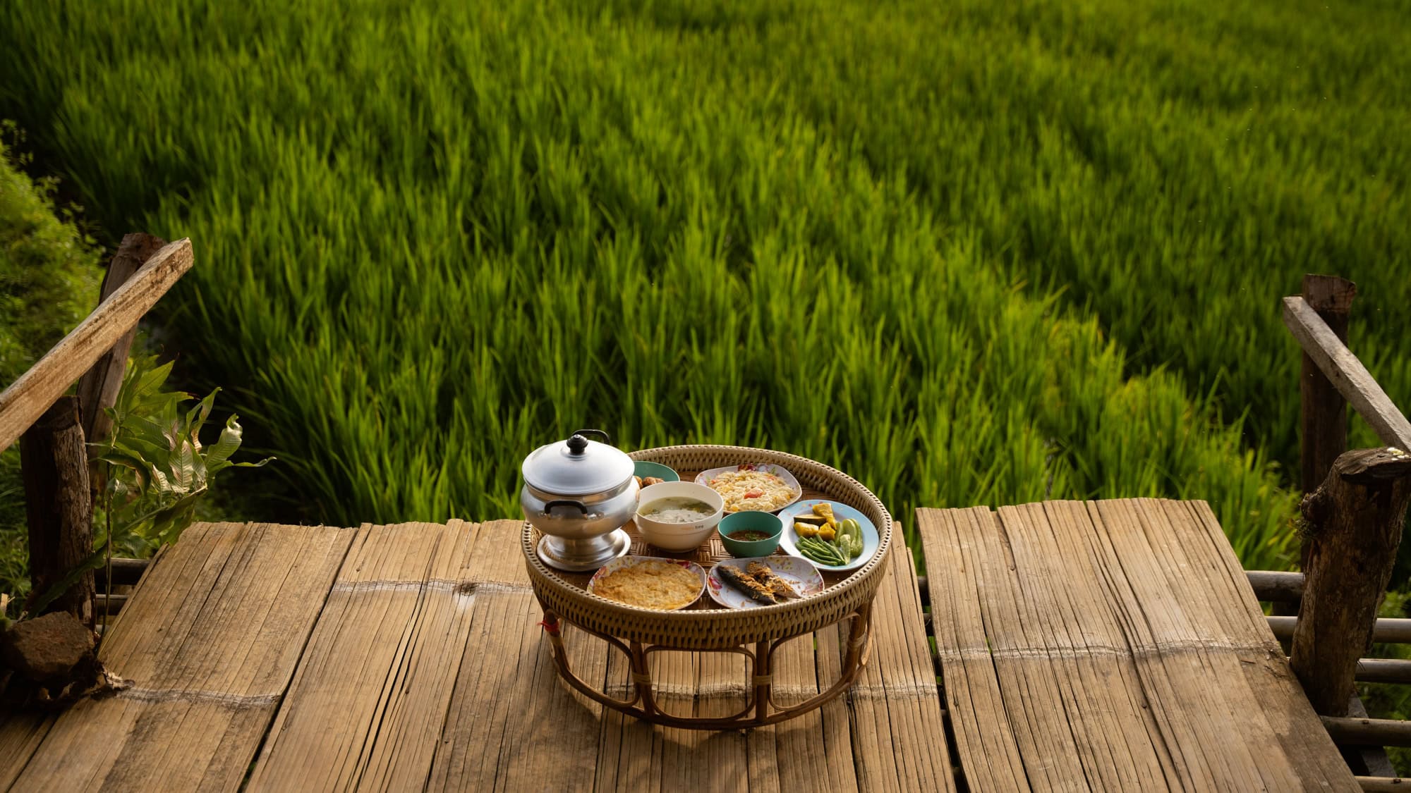 A woven tray filled with assorted dishes and rice sits on a wooden deck overlooking lush green rice paddies.