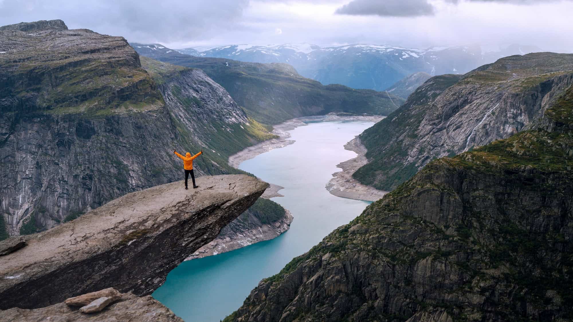 A hiker in an orange jacket stands triumphantly on Trolltunga, a dramatic rock ledge jutting over a fjord in Norway, surrounded by towering cliffs and a winding turquoise river below.
