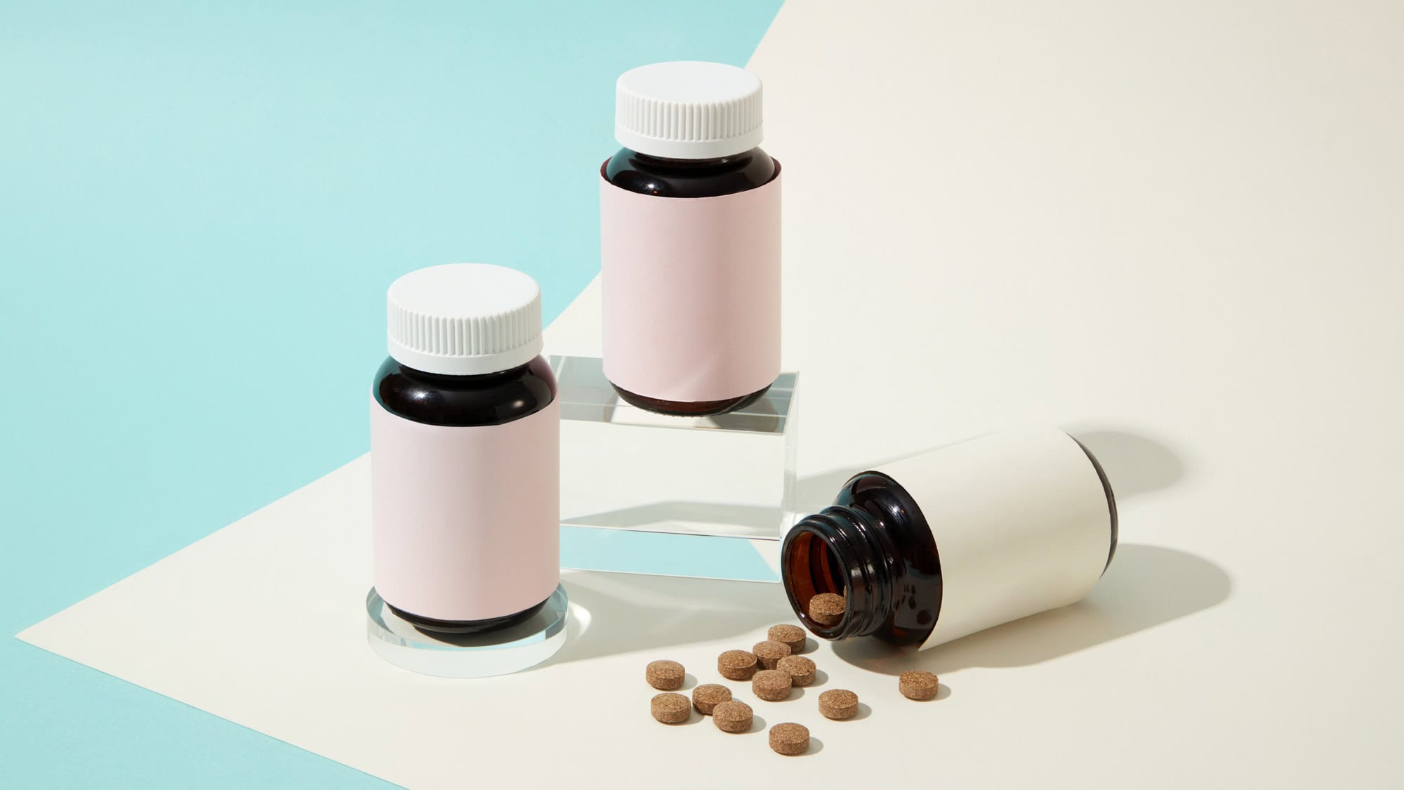 Three dark amber pill bottles with blank pastel labels sit on a color-blocked background of soft teal and cream, with one bottle tipped over, spilling round beige tablets.