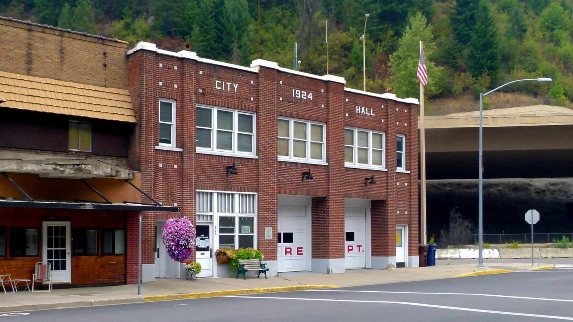 A historic red brick building marked "City Hall 1924" in white block lettering, located in Wallace, Idaho, with a U.S. flag flying beside it and lush green trees on the hillside behind.
