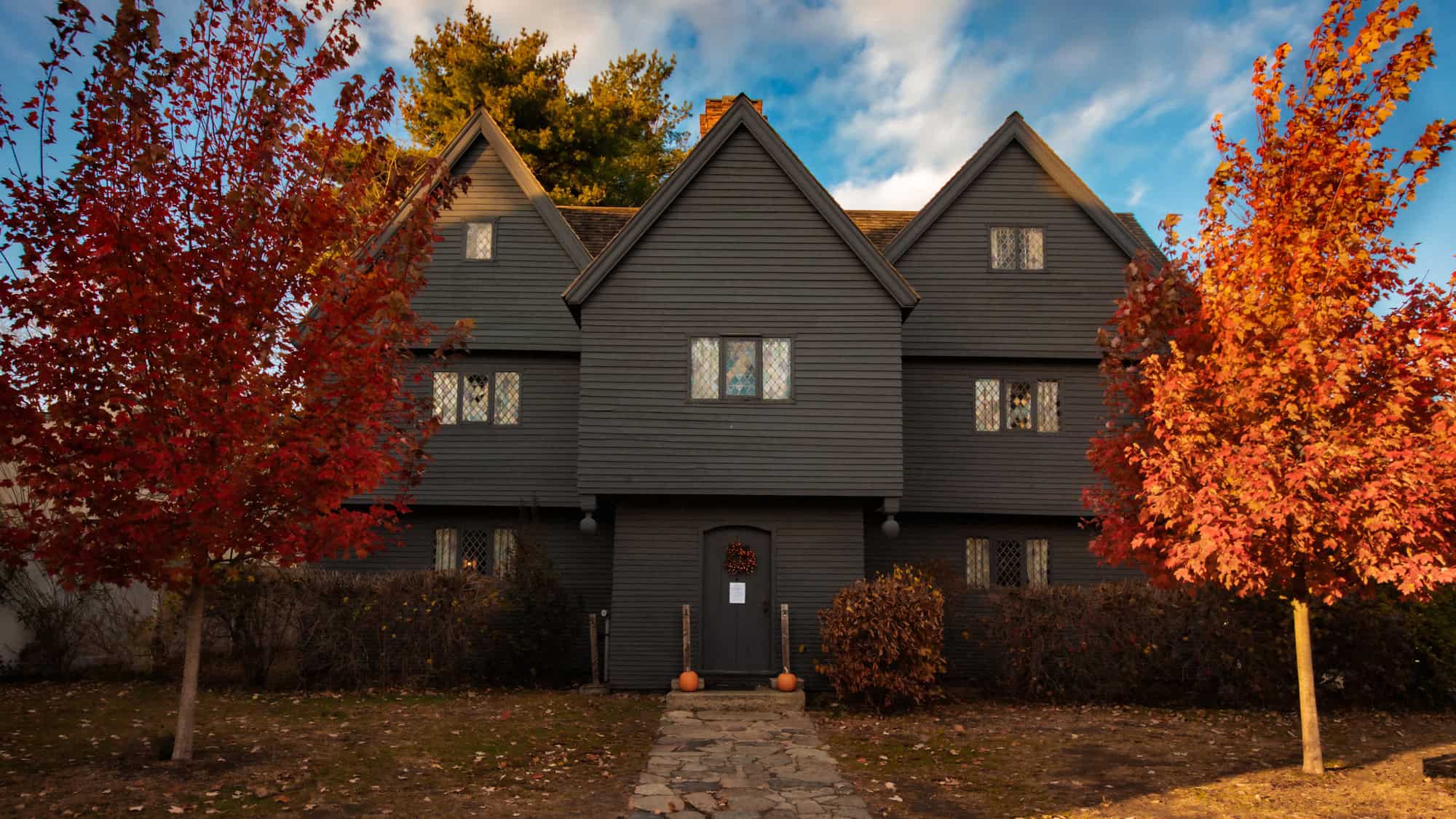 A dark gray, timber-framed colonial house with steep gables and diamond-pane windows, framed by vivid red-orange autumn trees under a partly cloudy sky—this is famously known as the Witch House, tied to the Salem witch trials.