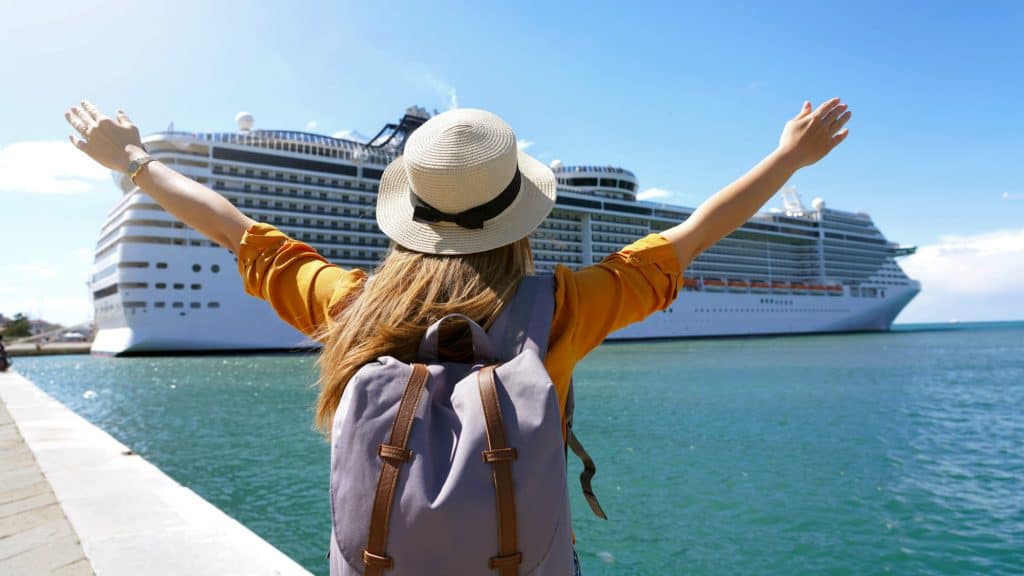 A woman with a straw hat and backpack stands with arms raised in excitement, facing a massive white cruise ship docked at port on a sunny day.