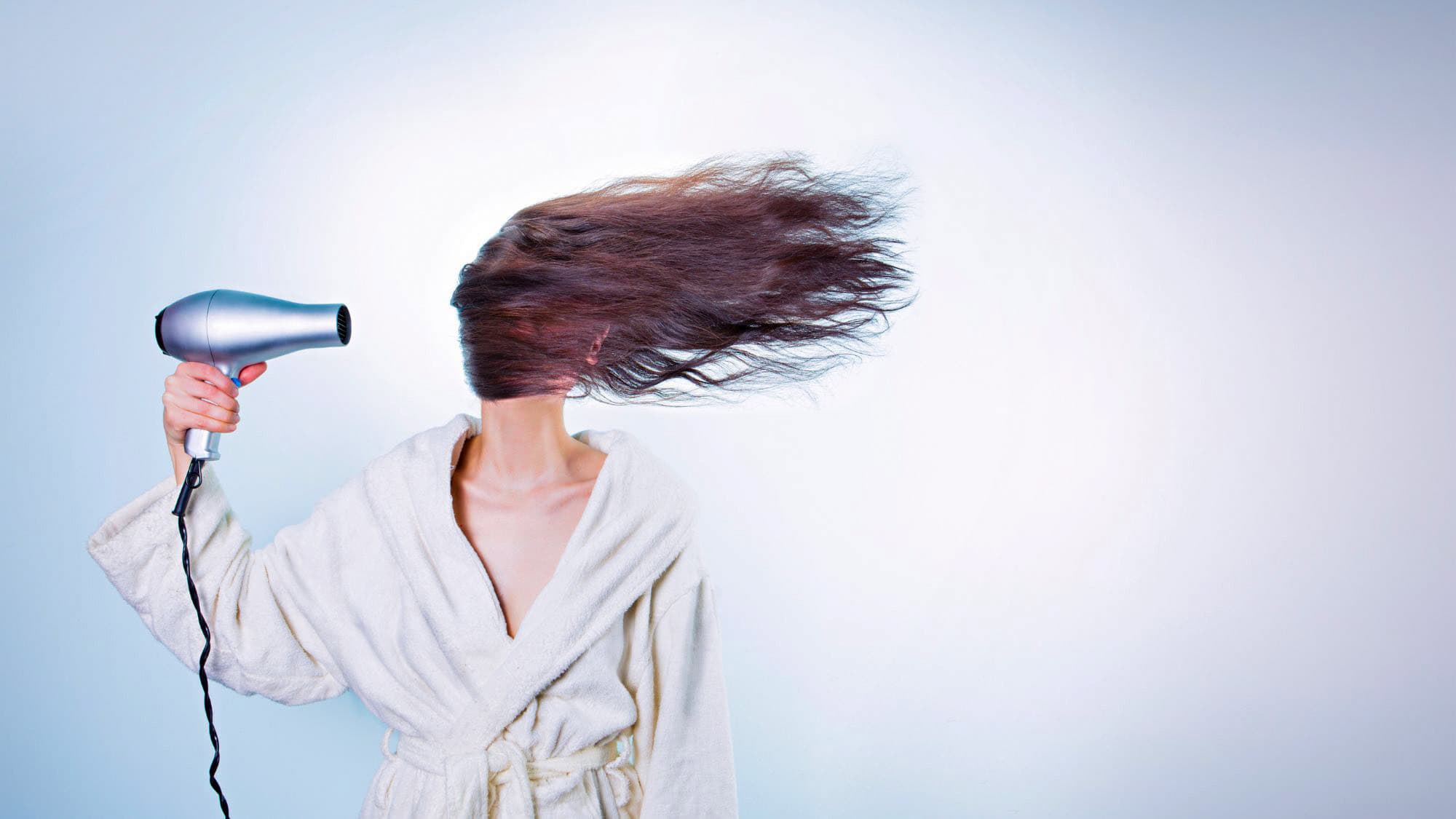 A woman in a white bathrobe blows her hair with a silver hairdryer, her long brown hair whipping dramatically across her face against a blue gradient background.