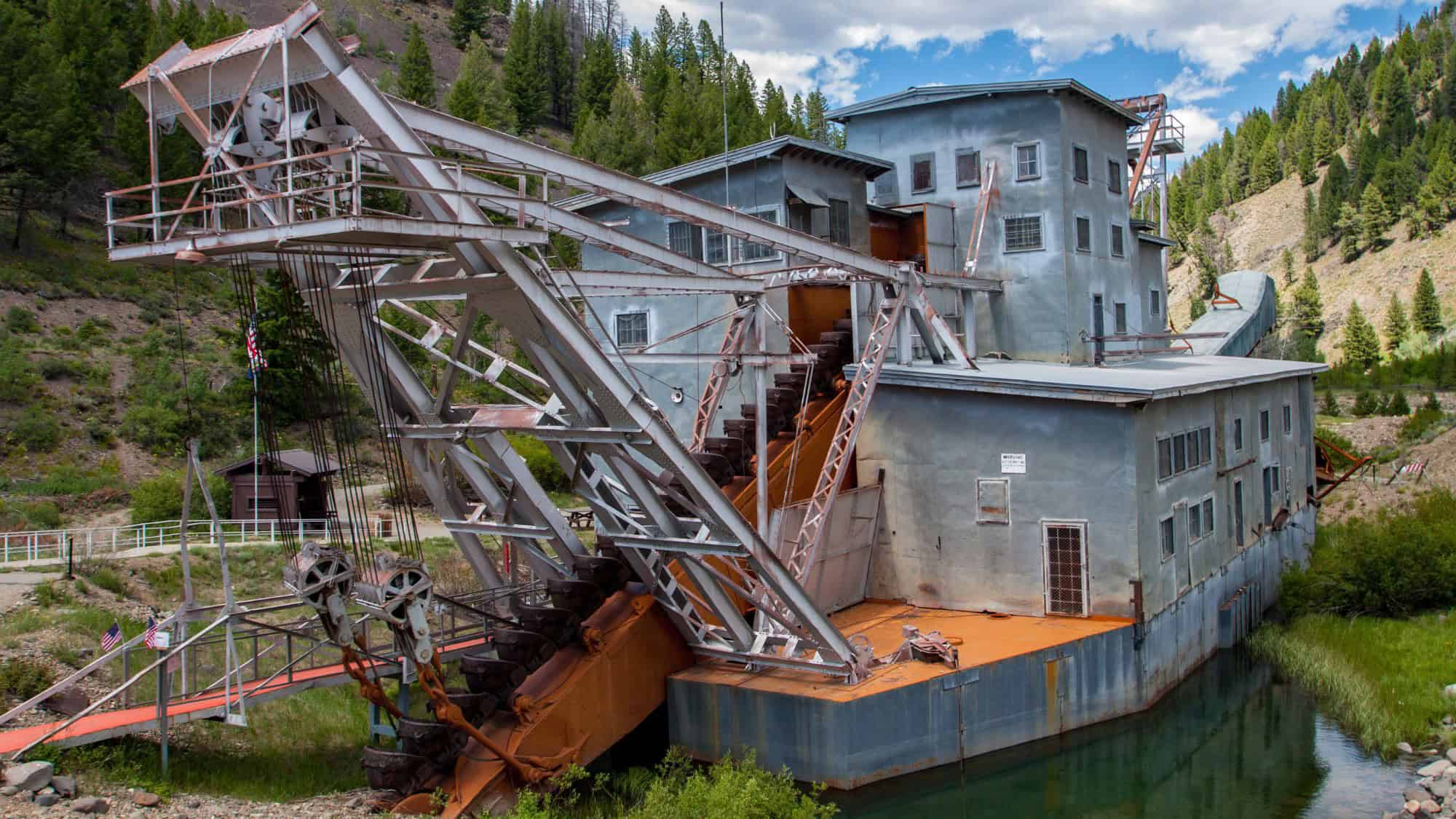 An old mining dredge with rusted components and towering metal arms, sitting in a narrow water channel surrounded by pine forest in a mountainous area.