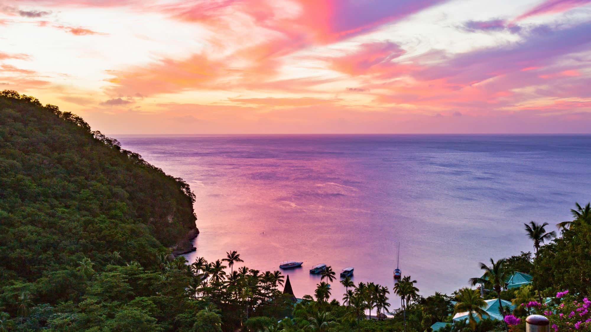 Vibrant sunset over Anse Chastanet Beach in St. Lucia, with hues of pink, orange, and purple reflecting on calm Caribbean waters, bordered by lush green hills, palm trees, and anchored boats near the shore.