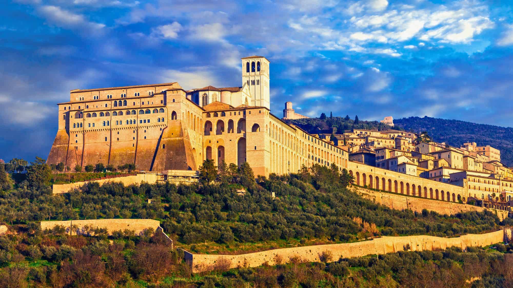 A grand hillside view of the Basilica of St. Francis of Assisi, showcasing its Romanesque architecture with arched arcades and a white bell tower, glowing in golden sunlight against a backdrop of dramatic clouds and rolling Umbrian hills.